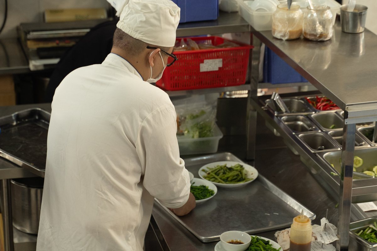 Một người thợ lớn tuổi mặc áo bếp trắng đứng tại quầy ra món, sắp rau và phần ăn lên khay